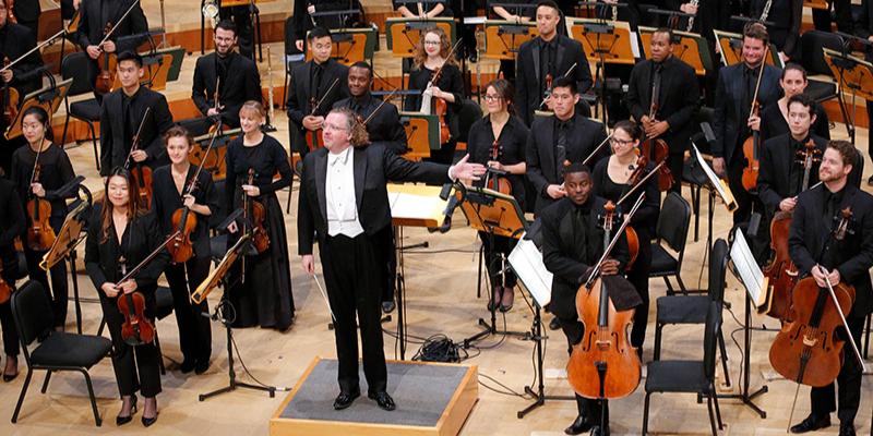 Conductor Stéphane Denève and the New World Symphony standing on stage facing the audience during applause.
